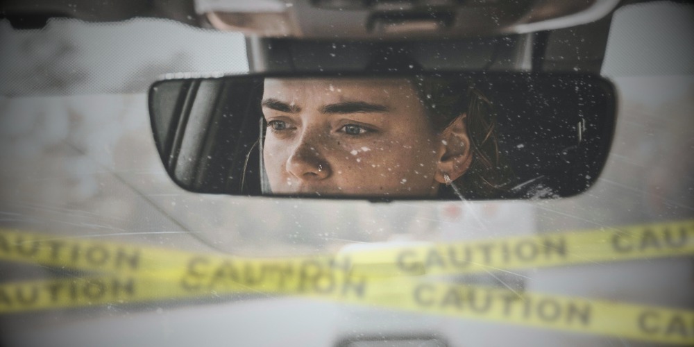 View of a rear-view mirror and front windshield. We can see the driver's worried-looking eyes looking at the road. In the blurry foreground is caution-tape covering her path.
