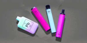 Four colorful e-cigarettes in various shapes are pictured on top of a cloudy gray background. Instead of brand names, the cigarettes are labeled with chemical element symbols for lead, etc.